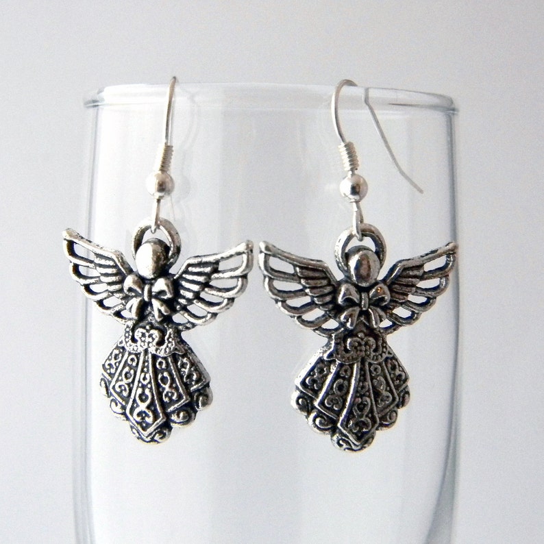 May include: Silver angel dangle earrings with intricate details and a bow on the chest. The earrings are hanging from a glass surface.