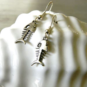 May include: A pair of silver fish bone earrings with a hook closure. The earrings are hanging from a white surface.