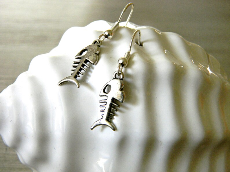 May include: A pair of silver fish bone earrings with a hook closure. The earrings are hanging from a white surface.