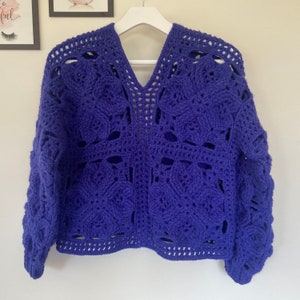 May include: A purple crocheted sweater with a V-neck and long sleeves. The sweater has a lacy pattern and a loose fit.