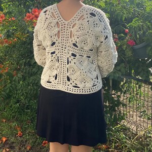 May include: A white crocheted sweater with a square pattern and a V-neck. The sweater has a loose fit and is worn over a black skirt.