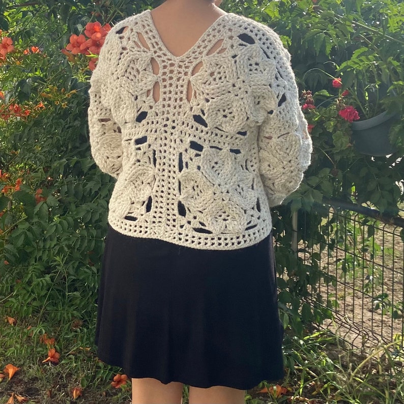 May include: A white crocheted sweater with a square pattern and a V-neck. The sweater has a loose fit and is worn over a black skirt.