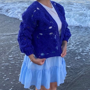 May include: A blue crocheted cardigan with a loose fit and open front. The cardigan has a textured design with large holes throughout. The cardigan is worn over a white t-shirt and a light blue skirt.