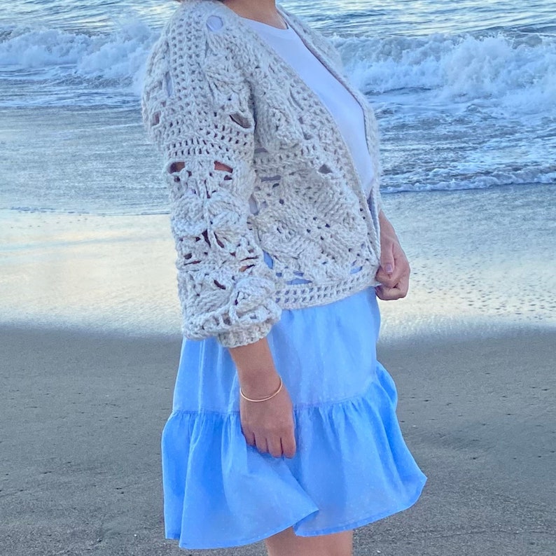 May include: A white crocheted cardigan with a delicate openwork pattern, worn over a white t-shirt and a light blue ruffled skirt. The person is standing on a sandy beach with ocean waves in the background.