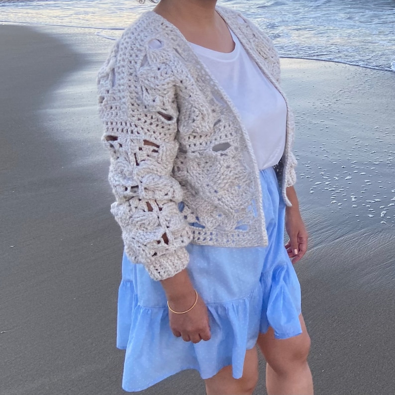 May include: A white crocheted cardigan with a delicate openwork pattern, worn over a white t-shirt and a light blue ruffled skirt. The person is standing on a sandy beach.