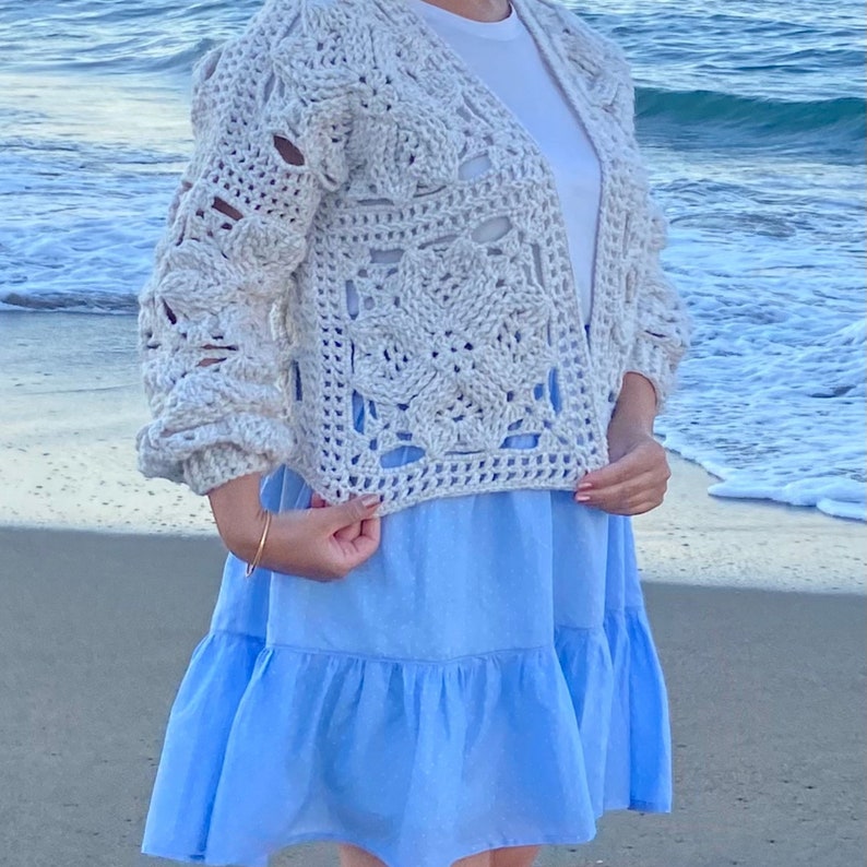 May include: A crocheted cardigan with a lacy pattern in a light beige color. The cardigan is open in the front and has long sleeves. The model is wearing the cardigan over a white t-shirt and a light blue dress.