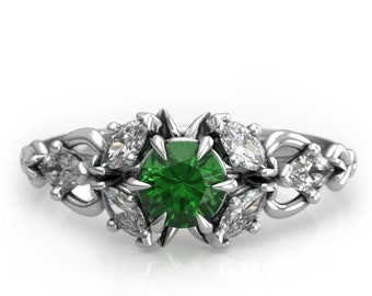 Könnte beinhalten: Ein silberner Ring mit einem grünen Edelstein, der von kleineren, klaren Edelsteinen umgeben ist. Der Ring hat ein zartes, filigranes Design.