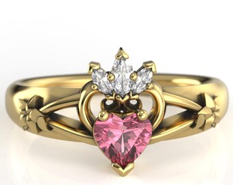 May include: A gold-colored ring featuring a pink heart-shaped gemstone at the center. The ring has a crown design above the heart and floral accents on the band. The ring is set against a white background.