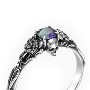 May include: A silver ring featuring a skull design and a large, iridescent purple gemstone. The ring is embellished with floral details.