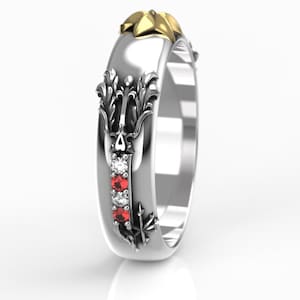 May include: A silver ring with a gold accent and ornate detailing. The ring features three red gemstones and two clear gemstones set in a vertical row. The design includes floral and leaf motifs.