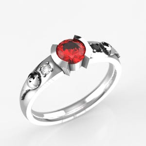 May include: A silver ring with a red gemstone in the centre and two smaller black and white gemstones on the band.