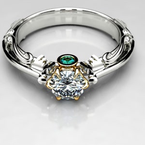 May include: A silver and gold engagement ring with a large, round, clear diamond center stone and a smaller, green emerald stone set above it. The ring has a detailed, ornate design.