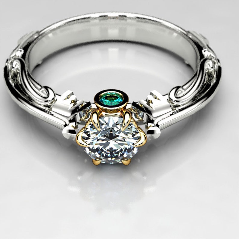 May include: A silver and gold engagement ring with a large, round, clear diamond center stone and a smaller, green emerald stone set above it. The ring has a detailed, ornate design.