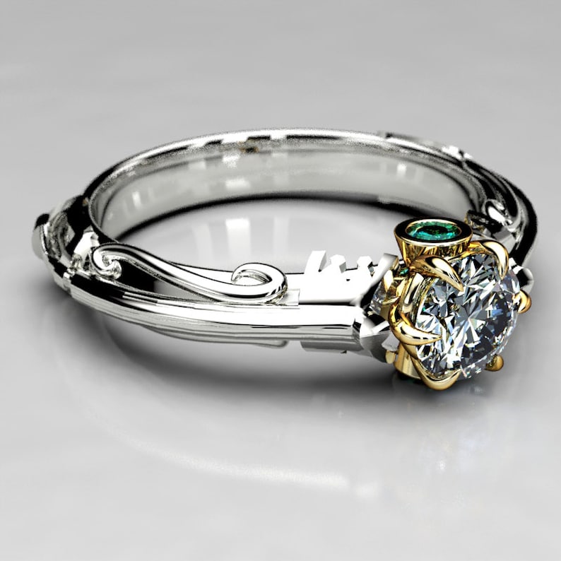 May include: A silver and gold engagement ring with a large, round, clear diamond and a small, green emerald stone. The ring has a delicate, ornate design.
