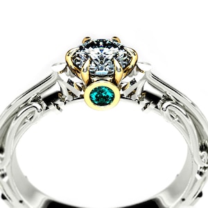 May include: A silver and gold engagement ring with a large, clear diamond center stone and a smaller, emerald green gemstone on the band.
