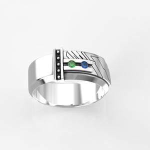 Puede incluir: Un anillo de plata con una banda texturizada y dos piedras preciosas. La piedra preciosa más grande es verde y la más pequeña es azul.