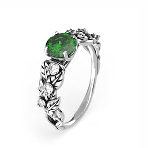 Puede incluir: Un anillo de plata con una piedra ovalada verde rodeada de piedras pequeñas transparentes. El anillo tiene un diseño de hojas.