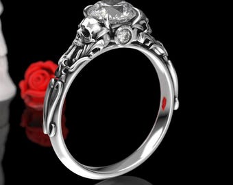 May include: A silver ring with a large, clear gemstone surrounded by two skull motifs. The ring is set against a black background.