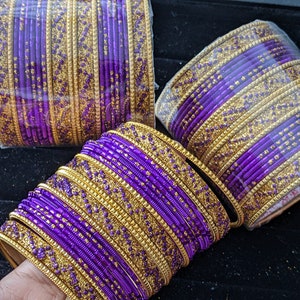 May include: Three stacks of purple and gold bangles. The bangles are a vibrant purple with gold embellishments. The gold accents are concentrated on the edges of the bangles. The bangles are stacked together, creating a visually appealing display.