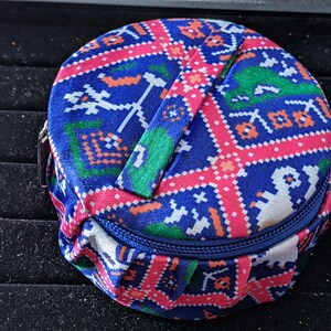 May include: A circular fabric pouch with a zip. The pouch has a colourful pattern of blue, pink, green, and orange geometric shapes. A matching fabric handle is attached. The pouch is likely for storing small items, such as jewellery.