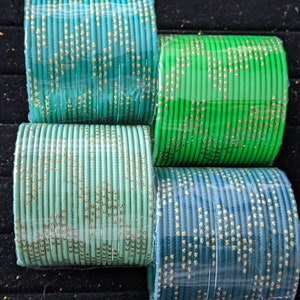 May include: An overhead shot showcases four bundles of colorful bangles. The bangles are in shades of teal, green, and blue, each adorned with a gold dotted pattern. The bangles are neatly arranged, creating a visually appealing display.