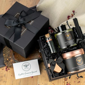 May include: A black gift box with a black ribbon and bow. The box contains a candle, diffuser, and a small bottle of essential oil. The box is sitting on a wooden surface with rose petals scattered around it. The candle and diffuser are both labelled "English Pear & Freesia".