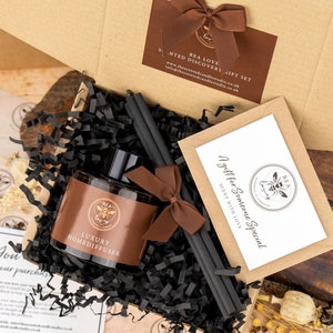 May include: A brown gift box filled with black shredded paper, a brown glass bottle with a black label that says "Luxury Homediffuser", black diffuser sticks, and a brown card with the text "A gift for Someone Special Scent with Love" and a bee logo.