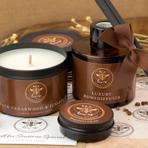 May include: A gift set featuring a brown candle with a black lid, a brown glass bottle with a black lid and brown ribbon, a small glass jar with matches, and a small tin with a brown lid. The candle and diffuser are labelled "Black Cedarwood & Juniper" and "Luxury Homediffuser" respectively. The label on the tin reads "BEA Loves".