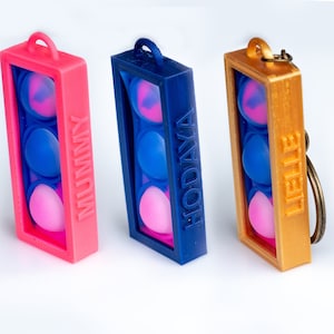 May include: Three keychain pop-it fidget toys. The first is pink with the word "NUMMY" on the side. The second is blue with the word "HODAYA" on the side. The third is gold with the word "HEILE" on the side.