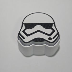 May include: A white and black Stormtrooper helmet design. The top of the helmet is white with a black, glittery visor. The bottom of the helmet is white with a scalloped edge. The design is a Star Wars theme.