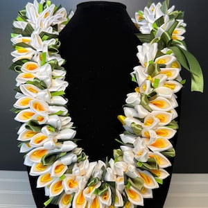 May include: A handcrafted lei necklace featuring white, yellow, and green satin ribbon flowers. The lei is displayed on a black mannequin head. The flowers are arranged in a layered pattern, creating a full and vibrant look.