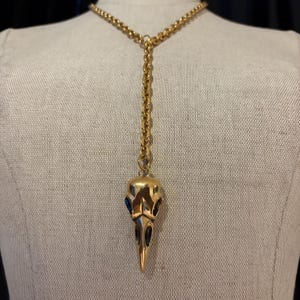 Könnte beinhalten: Eine goldene Kettenkette mit einem Vogel Schädel Anhänger. Der Schädel hat schwarze Details und blaue Augen.