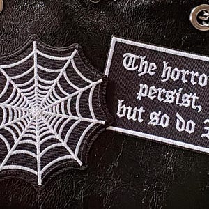 Könnte beinhalten: Zwei gestickte Aufnäher auf schwarzem Untergrund. Ein Aufnäher ist ein weißes Spinnennetz auf schwarzem Hintergrund. Der andere Aufnäher ist rechteckig mit dem Text "The horrors persist, but so do I" in Weiß.