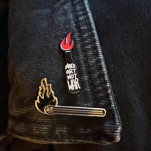 Könnte beinhalten: Zwei Emaille-Anstecknadeln auf einer Jeansjacke. Eine Nadel ist ein Streichholz mit einer Flamme, die andere ist eine schwarze Flasche mit roter Flamme und den Worten "MAKE ART NOT WAR" in Weiß. Die Anstecknadeln sind klein und detailliert.