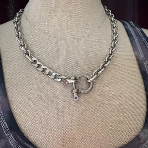 Op de afbeelding: Een zilveren ketting met een grote zilveren sluiting. De ketting is dik en heeft een platte schakels ontwerp.