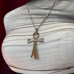 Könnte beinhalten: Halskette mit silberfarbenem Ankh-Anhänger. Das Ankh, ein Symbol des Lebens, hängt an einer feinen Kette. Der Anhänger hat eine polierte Oberfläche, die das Licht reflektiert. Die Halskette wird vor einem weiß-grauen Hintergrund präsentiert.