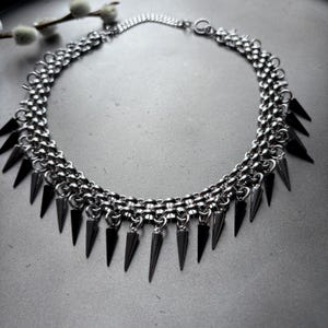 Könnte beinhalten: Eine silberne Kettenkette mit einer Reihe von scharfen, schwarzen Metallspitzen, die am unteren Ende hängen.