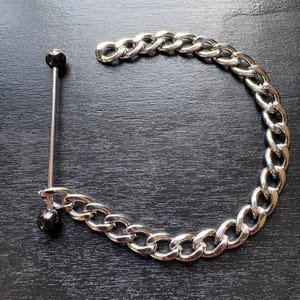 Könnte beinhalten: Eine silberfarbene Halskette mit großen Gliedern und einem Piercing im Langhantelstil. Die Langhantel hat einen langen, dünnen, silbernen Stab mit einer schwarzen Kugel an jedem Ende. Die Kette ist gebogen, und der Schmuck liegt auf einer dunklen Oberfläche.