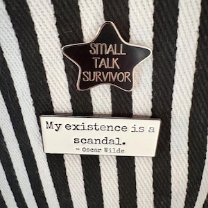 Könnte beinhalten: Zwei Emaille-Anstecknadeln auf einem schwarz-weiß gestreiften Hintergrund. Die obere Anstecknadel ist ein schwarzer Stern mit den Worten "SMALL TALK SURVIVOR". Die untere Anstecknadel ist rechteckig und trägt die Aufschrift "My existence is a scandal. - Oscar Wilde."