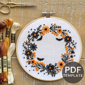 May include: A Halloween-themed embroidery hoop with a floral wreath design in black and orange thread. The wreath includes bats, spiderwebs, and flowers. The image also shows embroidery floss and gold scissors. The text "PDF TEMPLATE" is visible.