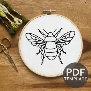 May include: A black and white line drawing of a bee on white fabric in an embroidery hoop. The bee has wings, a body with stripes, and six legs. The hoop is on a wooden surface.