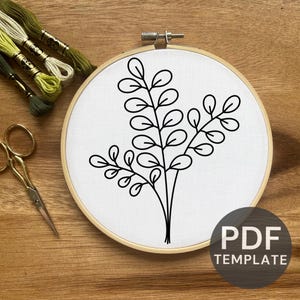 May include: Embroidery hoop with a botanical design. The design features a black outline of leaves and stems on white fabric. The hoop is made of wood. The image also includes embroidery floss and scissors.