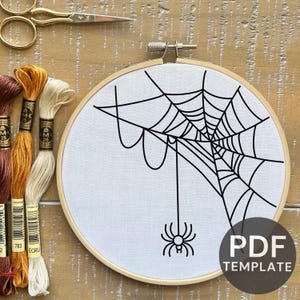 Może przedstawiać: Okrągła tamborek z białym materiałem z czarnym wzorem pajęczyny i wiszącym pająkiem. Obraz zawiera nici do haftu w odcieniach brązu, złota i kremu, a także złote nożyczki. Widoczny jest również szablon PDF.