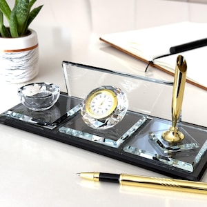 May include: A rectangular glass desk set featuring a clear glass base, a small crystal bowl, a round clock with a gold-tone bezel and white face, and a gold-tone pen holder. A matching gold-tone pen with a black grip rests in front of the set.