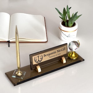 Puede incluir: Una placa de nombre de escritorio marrón con un portaplumas dorado y un pequeño globo terráqueo. La placa tiene el texto "Benjamin McGill, Attorney at Law" y un icono dorado de balanza de la justicia.