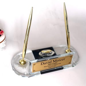 May include: A clear glass pen holder with two gold pens. The holder has a gold nameplate that reads "David Mercer, Store Leader, Tom James of Chattanooga, August 2023". A small inkwell is also included.