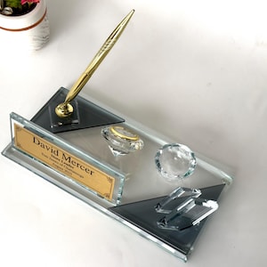 May include: A rectangular glass desk set featuring a gold pen in a holder, two clear crystal-like decorative accents, and a gold-toned nameplate engraved with the text David Mercer, Store Leader, Tom James of Chattanooga, August 2023.