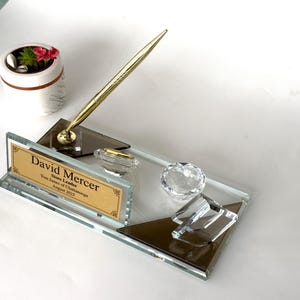 May include: A glass desk nameplate featuring a gold-toned pen in a holder, a clear crystal accent, and a gold plaque engraved with the text David Mercer, Store Leader, Tom James of Chattanooga, August 2025, set on a rectangular glass base.