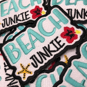 May include: A close-up of several embroidered patches with the text "Beach Junkie" in turquoise blue lettering on a white background. The patches have a black border and feature a yellow starfish and a red hibiscus flower.