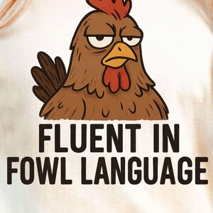 Könnte beinhalten: Cremefarbenes T-Shirt mit einem Cartoon-Huhn mit grimmigem Gesichtsausdruck. Das Huhn ist braun mit rotem Kamm und Kehllappen. Der Text "FLUENT IN FOWL LANGUAGE" ist darunter in Schwarz gedruckt.
